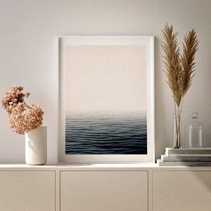 May include: A framed print of a seascape with a calm, dark blue ocean meeting a pale, hazy sky. The print is displayed on a light-colored cabinet, next to a white vase with dried flowers and a glass vase with tall, dried grasses.
