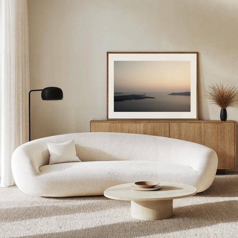 May include: A curved, cream-coloured sofa with a single square throw pillow, positioned in front of a wooden sideboard. A low, oval-shaped coffee table sits in the foreground, and a framed landscape print hangs on the wall above the sideboard.