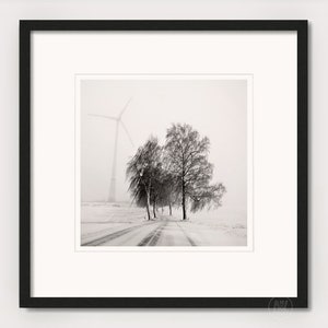 May include: A black and white photograph of a snowy road with trees lining the sides. A windmill is visible in the distance, partially obscured by fog.