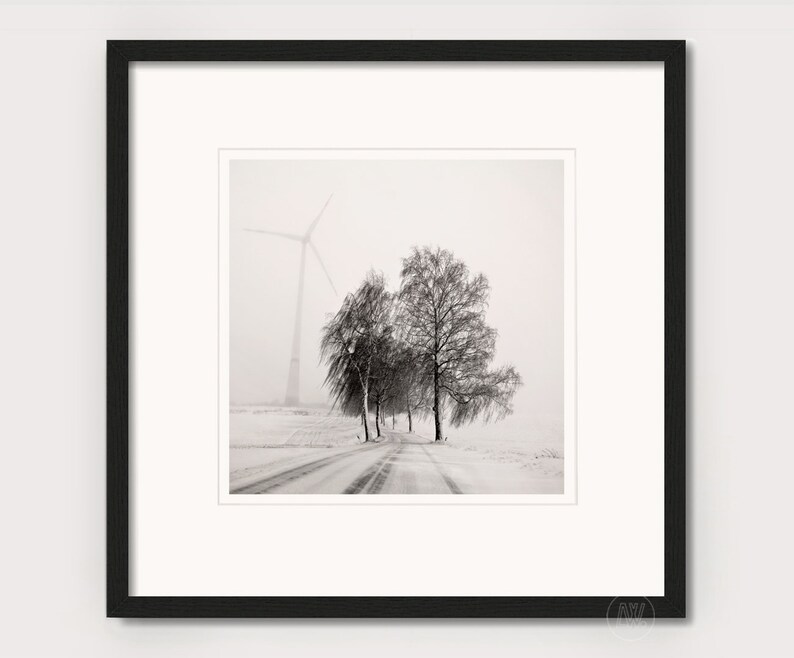 May include: A black and white photograph of a snowy road with trees lining the sides. A windmill is visible in the distance, partially obscured by fog.