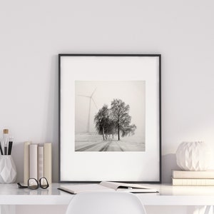 May include: A black and white photograph of a snowy landscape with a windmill in the distance and a few trees in the foreground. The photo is framed in a black frame and is hanging on a white wall.