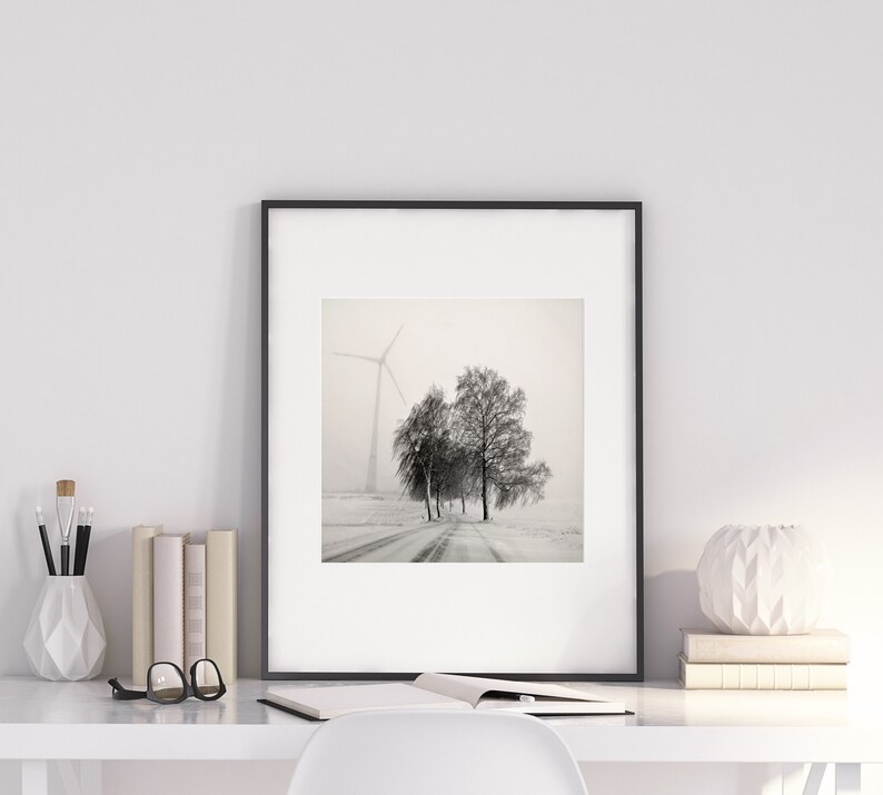 May include: A black and white photograph of a snowy landscape with a windmill in the distance and a few trees in the foreground. The photo is framed in a black frame and is hanging on a white wall.