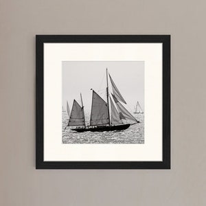 May include: A black and white photograph of a sailboat with its sails billowing in the wind. The sailboat is in the foreground of the image, with other sailboats in the background.