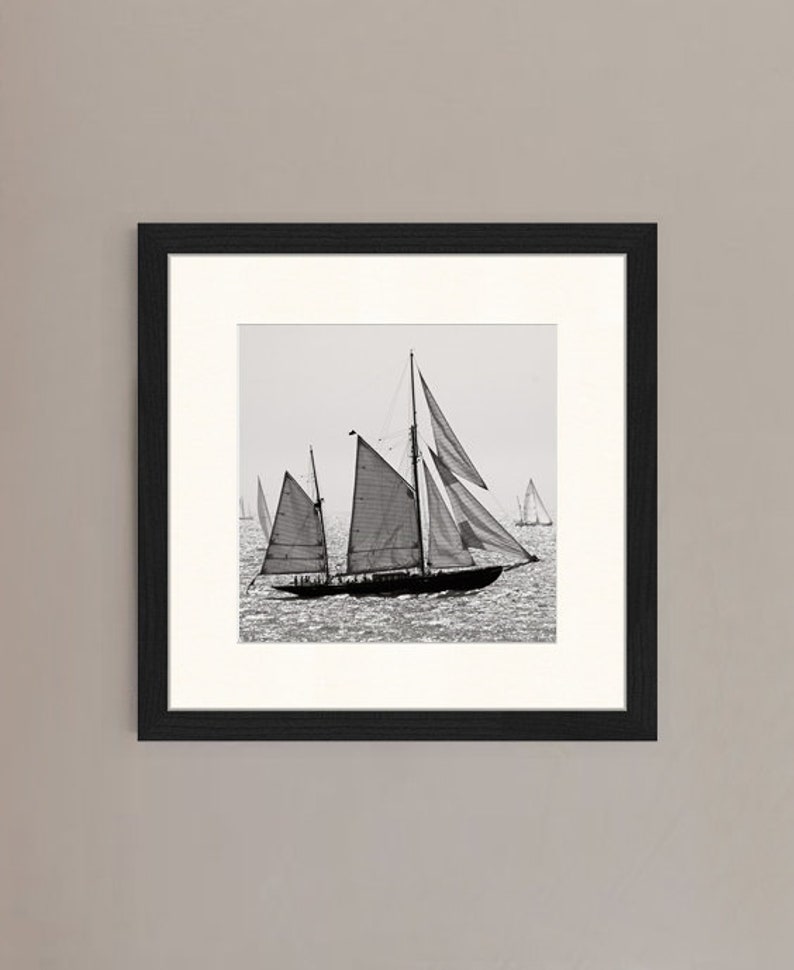 May include: A black and white photograph of a sailboat with its sails billowing in the wind. The sailboat is in the foreground of the image, with other sailboats in the background.