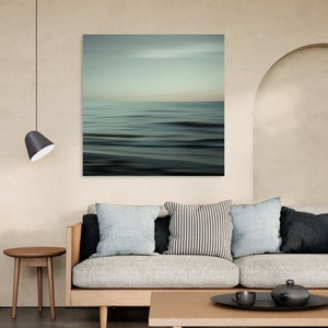 May include: A light grey sofa with three cushions, a black and white striped cushion, a light blue cushion, and a dark grey cushion. The sofa is in front of a wall with a large abstract painting of a blue and green ocean scene. A small wooden table with a cup and a teapot is in front of the sofa.