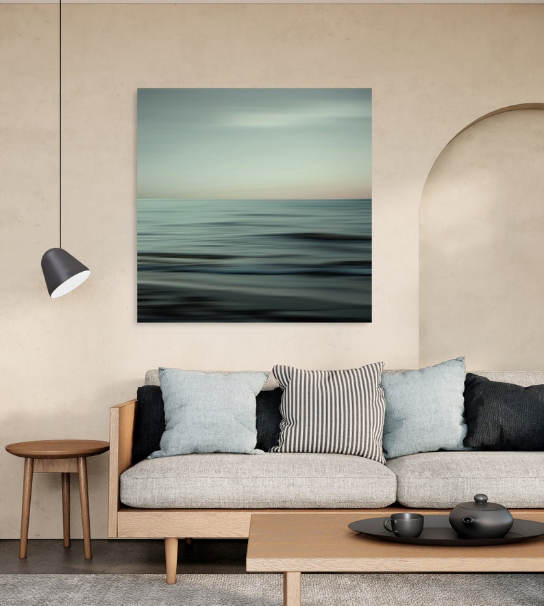 May include: A light grey sofa with three cushions, a black and white striped cushion, a light blue cushion, and a dark grey cushion. The sofa is in front of a wall with a large abstract painting of a blue and green ocean scene. A small wooden table with a cup and a teapot is in front of the sofa.