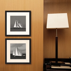 May include: Two black framed photographs of sailboats on a wooden wall. The top photo shows a sailboat with its sails full, while the bottom photo shows a sailboat with its sails partially furled.