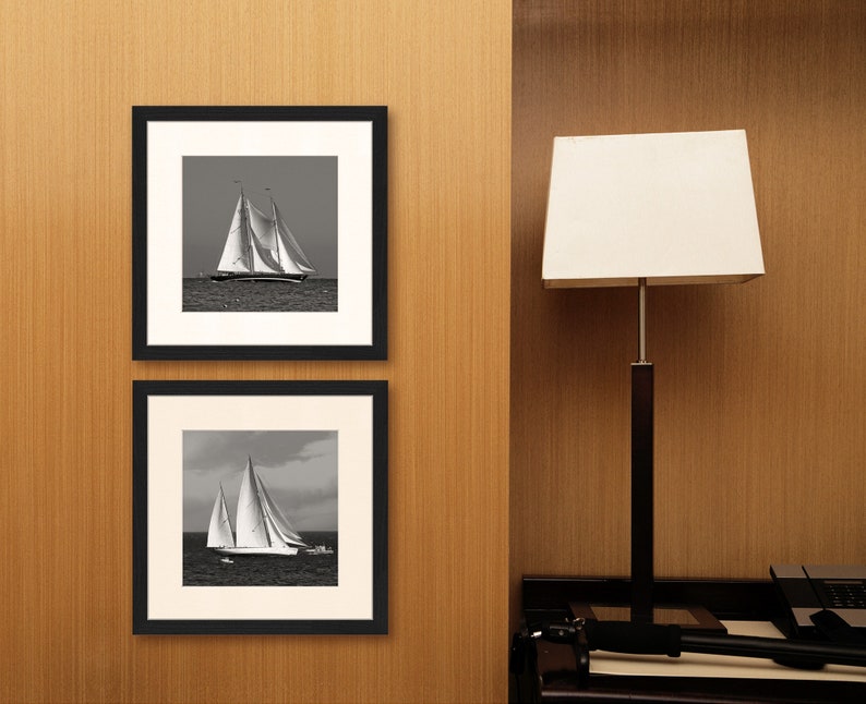May include: Two black framed photographs of sailboats on a wooden wall. The top photo shows a sailboat with its sails full, while the bottom photo shows a sailboat with its sails partially furled.
