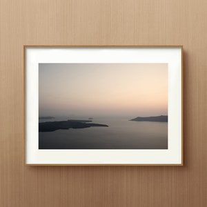 May include: A framed landscape photograph featuring a calm body of water surrounded by dark, silhouetted landmasses under a soft, pale gradient sky. The image is enclosed in a simple, light-toned wooden frame with a wide white mat border.