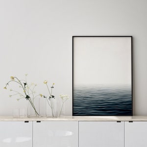 May include: A framed art print featuring a minimalist seascape with a gradient sky and dark blue water. The artwork is displayed on a white cabinet, accompanied by clear glass vases with delicate flowers.