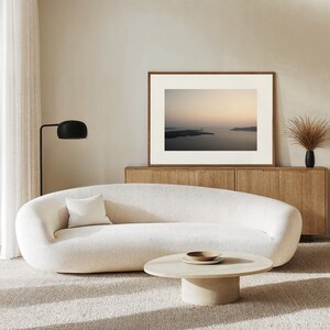 May include: A curved, cream-coloured sofa with a single square throw pillow, paired with a low, oval-shaped light wood coffee table. A framed landscape photograph hangs above a wooden sideboard, accompanied by a black floor lamp and dried grass.