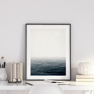 May include: A framed art print depicting a serene ocean scene with dark blue water and a misty horizon. The artwork is displayed on a white desk, alongside books, a vase with brushes, and a decorative white sphere.