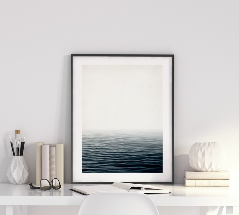 May include: A framed art print depicting a serene ocean scene with dark blue water and a misty horizon. The artwork is displayed on a white desk, alongside books, a vase with brushes, and a decorative white sphere.