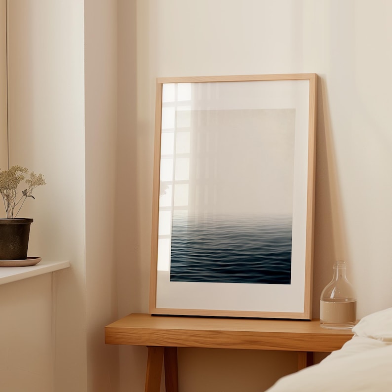 May include: A framed art print featuring a serene ocean scene. The artwork depicts dark blue water with gentle waves, fading into a misty, light-colored sky. The print is encased in a light-colored wooden frame, creating a minimalist aesthetic.