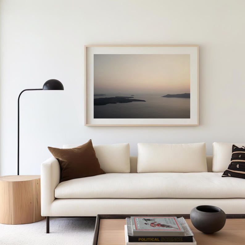 May include: A framed landscape photograph of a dark sea and distant islands at dusk, hanging above a cream-coloured sofa. The room includes a black floor lamp, a light wood cylindrical side table, and a coffee table with books and a dark bowl.