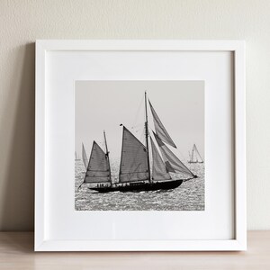 May include: A black and white photograph of a sailboat with its sails billowing in the wind. The sailboat is in the foreground, with other sailboats in the background.