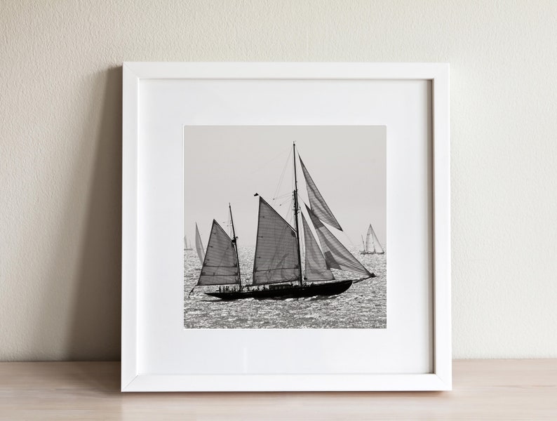 May include: A black and white photograph of a sailboat with its sails billowing in the wind. The sailboat is in the foreground, with other sailboats in the background.