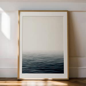 May include: A framed art print featuring a serene ocean scene. The artwork depicts a gradient of colors, transitioning from a light beige sky to deep blue water. The waves are subtly visible, creating a sense of calm. The frame is a light wood color.