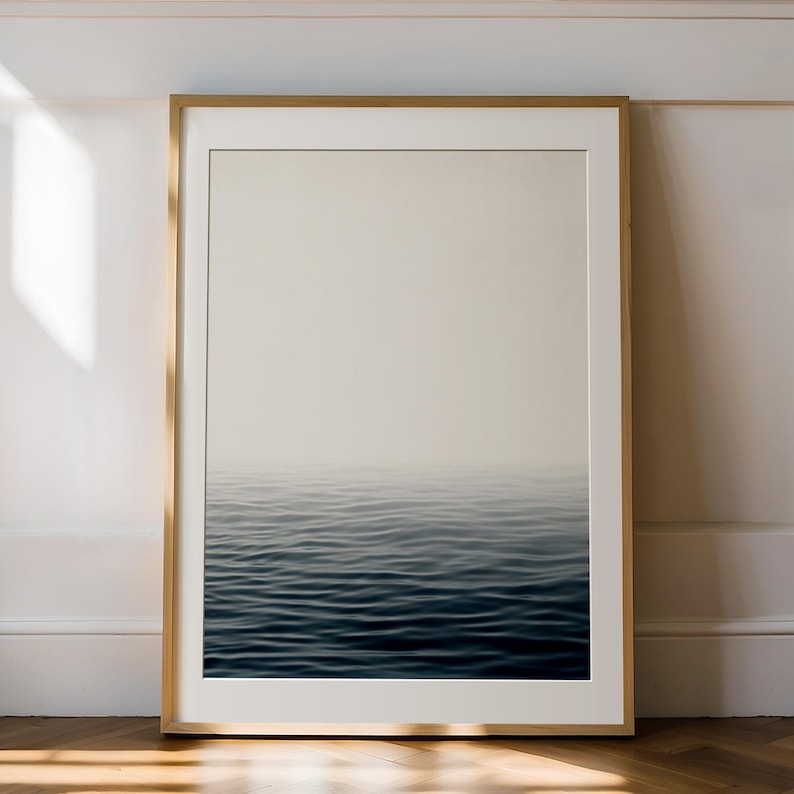 May include: A framed art print featuring a serene ocean scene. The artwork depicts a gradient of colors, transitioning from a light beige sky to deep blue water. The waves are subtly visible, creating a sense of calm. The frame is a light wood color.