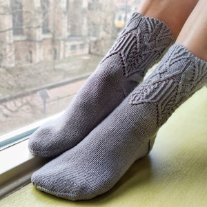 May include: A pair of grey knitted socks with a delicate lace pattern. The socks are worn on a person's feet and are resting on a yellow surface.