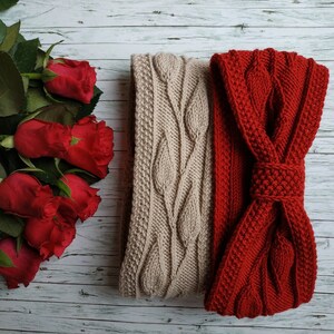 May include: Two knitted headbands, one beige with a cable knit pattern and one red with a bow. The headbands are on a white wooden background.