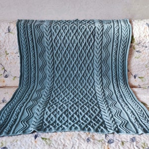 Op de afbeelding: Een tealgekleurde gebreide deken met een ingewikkeld kabelbreipatroon. De deken heeft een herhalend ruitpatroon in het midden, omzoomd door getextureerde verticale panelen en een geschulpte rand. De deken is over een bank met bloemenpatroon gedrapeerd.