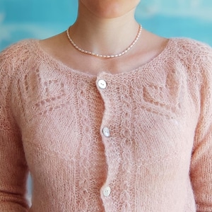 May include: A light pink, open-knit cardigan with a scoop neck and three white buttons. The cardigan has a delicate, lacy pattern on the upper chest and sleeves. A strand of small, light-colored beads is worn around the neck.