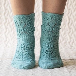 May include: A pair of light blue knitted socks with an intricate floral and geometric lace pattern. The socks are knee-high and feature a textured design with small, raised bobbles. The socks are displayed on a white, textured surface.