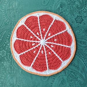 Puede incluir: Un posavasos de rodaja de pomelo tejido a crochet en tonos naranja, blanco y rosa. El posavasos está hecho con un diseño texturizado y tejido.