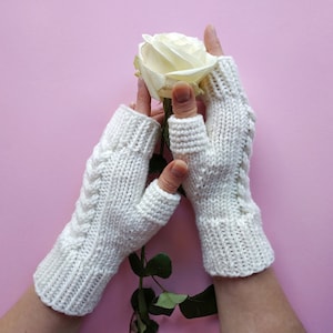 May include: A pair of white knitted fingerless gloves with a cable knit pattern. The gloves are held by a person who is also holding a white rose.