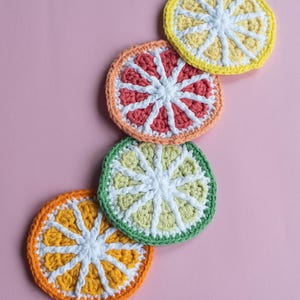 May include: Four crocheted citrus fruit coasters in shades of yellow, orange, red, and green. The coasters are arranged in a row on a pink background.