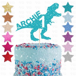 May include: A turquoise glitter dinosaur cake topper with the name "ARCHIE" and the number "1". The cake is light blue with colorful sprinkles. Several glitter star decorations are also visible.