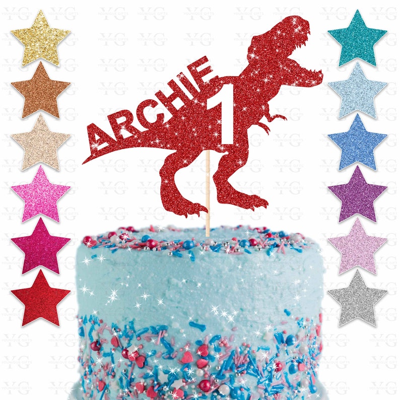 May include: A red glitter dinosaur cake topper with the name "ARCHIE" and the number "1". The topper is on a wooden stick and is placed on a blue frosted cake with colorful sprinkles. Glitter star decorations are on the left and right.