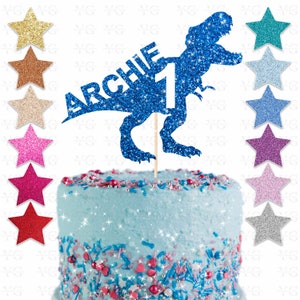 May include: A blue glitter dinosaur cake topper with the name "ARCHIE" and the number "1". The topper is on a light blue frosted cake with colorful sprinkles and star-shaped decorations.