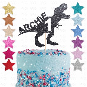 May include: A black glitter dinosaur cake topper with the name "ARCHIE" and the number "1". The topper is on a wooden stick and is placed on a blue frosted cake with colorful sprinkles and star-shaped decorations.