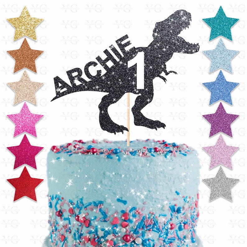 May include: A black glitter dinosaur cake topper with the name "ARCHIE" and the number "1". The topper is on a wooden stick and is placed on a blue frosted cake with colorful sprinkles and star-shaped decorations.
