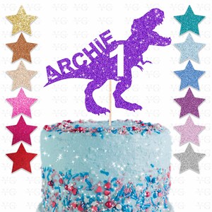 May include: A purple glitter dinosaur cake topper with the name "ARCHIE" and the number "1". The topper is on a light blue frosted cake with colorful sprinkles and star-shaped decorations.