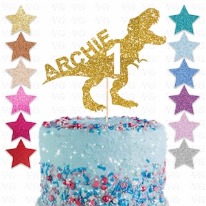 May include: A gold glitter cake topper in the shape of a Tyrannosaurus Rex with the name "ARCHIE" and the number "1". The topper is on a wooden stick and is placed on a blue frosted cake with colorful sprinkles. Several glitter star decorations are also visible.