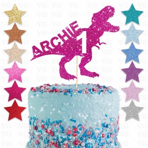 May include: A pink glitter cake topper featuring a Tyrannosaurus Rex silhouette with the name "ARCHIE" and the number "1". The topper is on a wooden stick, placed on a blue frosted cake with colorful sprinkles. Glitter star decorations are in the background.
