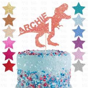 May include: A glittery orange Tyrannosaurus Rex cake topper with the name "ARCHIE" and the number "1". The cake is light blue with colorful sprinkles. Various glitter star decorations are arranged around the cake.
