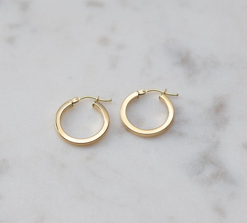 May include: A pair of small, polished gold hoop earrings with a smooth, flat surface and a hinged post closure. The earrings feature a circular design with a consistent thickness, showcasing a bright, reflective metallic finish throughout the piece.