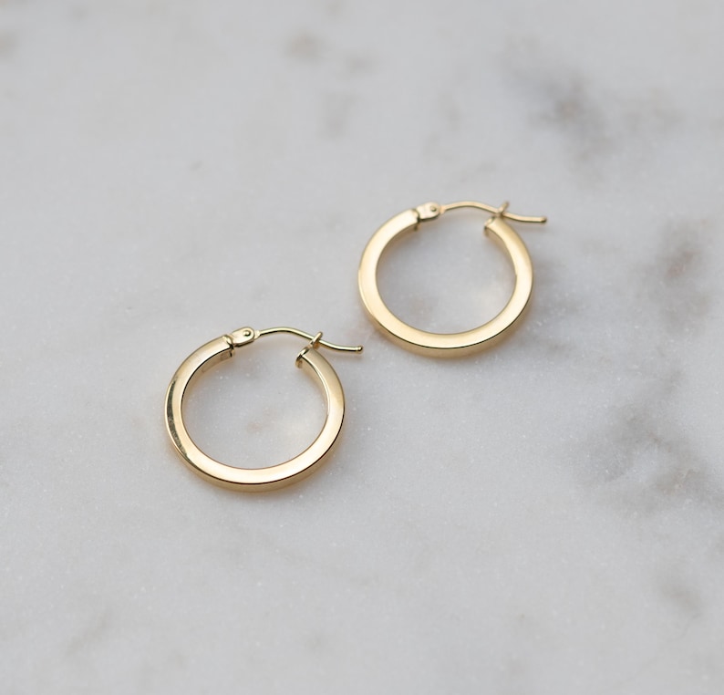 May include: A pair of small, polished gold hoop earrings with a smooth, rounded surface and a hinged post closure. The earrings feature a simple, circular design with a reflective metallic finish, showcasing a classic and clean aesthetic.