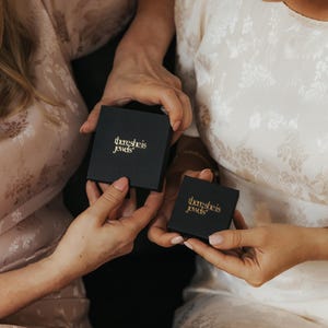 May include: Two square black jewelry boxes with the gold text there she is jewels printed on the lids. The boxes are held in place, with one larger box positioned above a smaller box, both featuring a matte finish and clean, sharp edges.