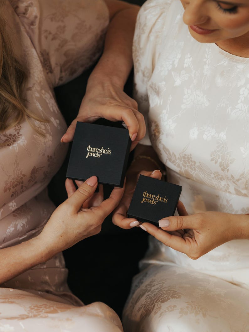 May include: Two square black jewelry boxes with the gold text there she is jewels printed on the lids. The boxes are held in place, with one larger box positioned above a smaller box, both featuring a matte finish and clean, sharp edges.