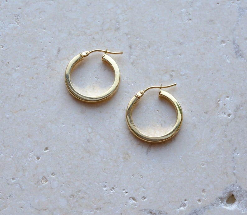 May include: A pair of polished gold hoop earrings with a smooth, rounded profile and a hinged post closure. The earrings feature a classic circular design with a reflective metallic finish, showcasing a simple and clean aesthetic for everyday wear.
