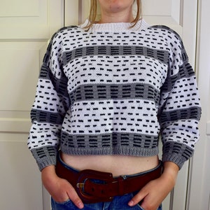 Könnte beinhalten: Ein kurzer grauer und weißer gestreifter Pullover mit einem schwarzen geometrischen Muster. Der Pullover hat lange Ärmel und einen runden Ausschnitt.