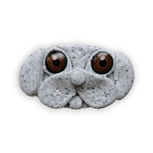 Könnte beinhalten: Hellgraue Hundegesicht-Skulptur mit gesprenkelter Textur und großen, braunen Augen. Die Skulptur hat eine markante Nase und Schlappohren, was ihr ein cartoonartiges Aussehen verleiht. Einzigartiges dekoratives Element.