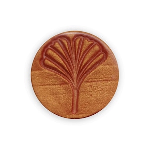 Könnte beinhalten: Ein rundes, metallisches goldfarbenes Siegelwachs mit einem erhabenen, roten Ginkgoblatt-Design.