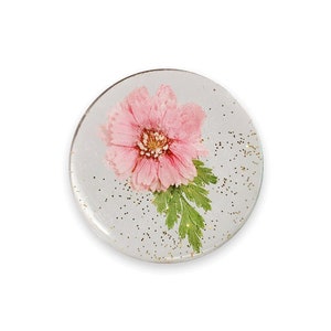 Könnte beinhalten: Ein runder, transparenter Harz-Untersetzer mit einer rosa Blume und grünen Blättern, die darin gepresst sind. Der Untersetzer hat einen goldenen Glitzer-Hintergrund.