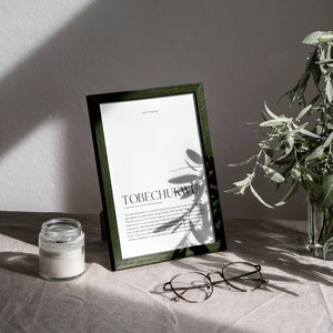 May include: A green framed print with the word "TOBECHUKWU" in black text. The print is on a white background and has a definition of the word below it. The print is sitting on a light brown surface with a white candle and round glasses.